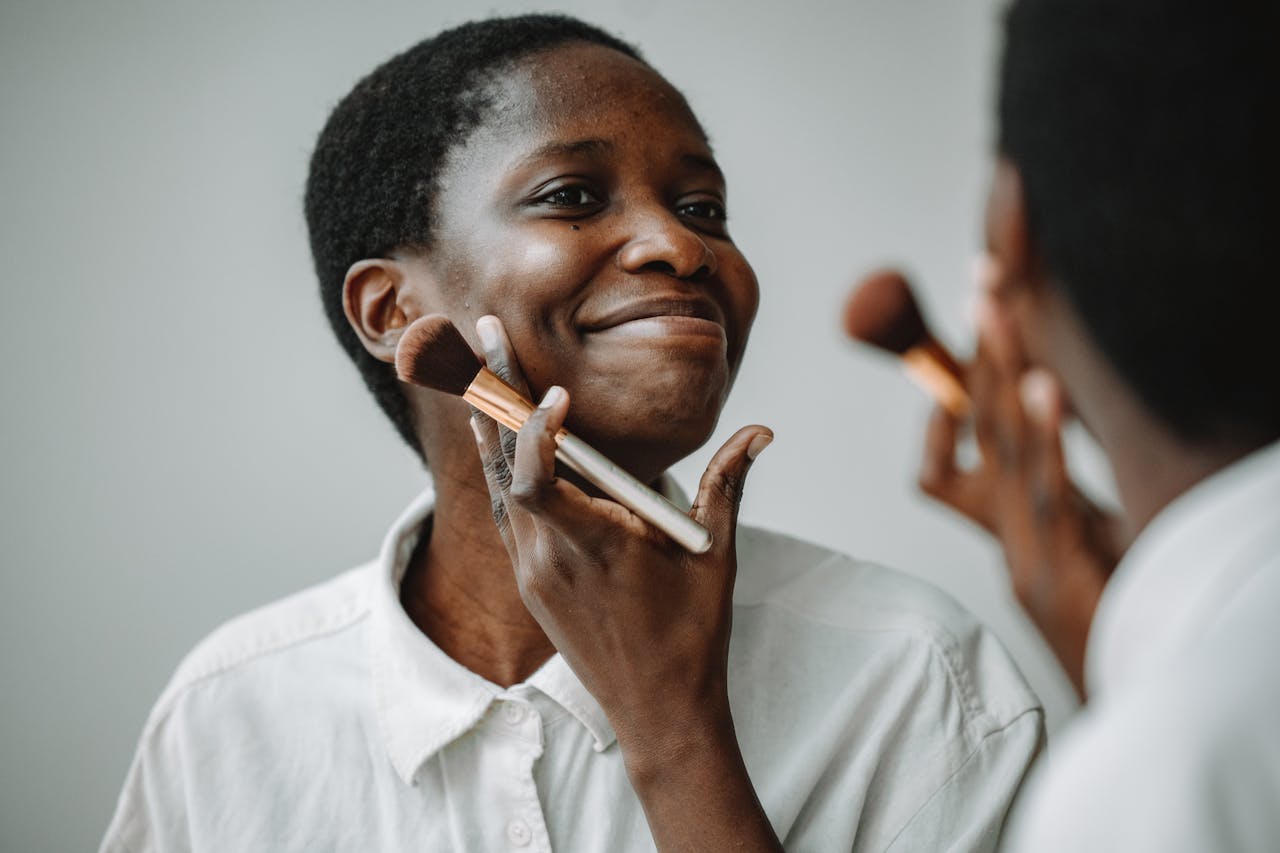 Smiling woman looking in the mirror while applying makeup with a brush indoors.